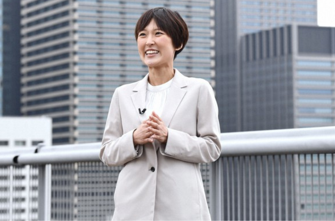 元NHKアナウンサーの近江友里恵さん。都市のビル群を背景に、晴れやかな笑顔でインタビューに応じている様子。